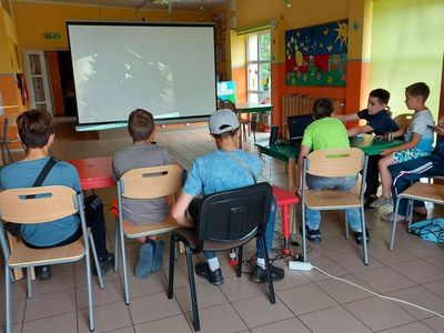 Wychowankowie z Placówki Wsparcia Dziennego w Laskach oglądają bajkę na dużym ekranie, ich plecy na pierwszym planie, na drugim planie ekran.