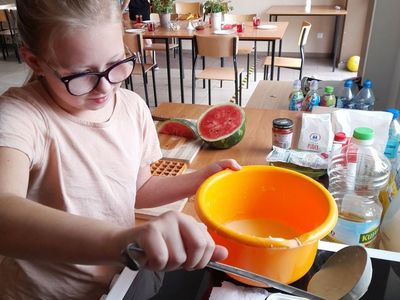 Dziewczynka z Placówki Wsparcia Dziennego w Sulechowie, wlewająca ciasto do gofrownicy. W tle dziewczynka stojąca przy biurku i stół przygotowany do posiłku.
