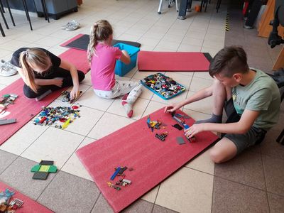 Dwie dziewczynki i jeden chłopiec z Placówki Wsparcia Dziennego w Sulechowie , siedzący na matach gimnastycznych i budujący z klocków Lego.