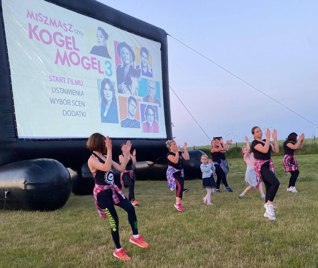 Grupa zumbowiczek tańczy zumbę na boisku rekreacyjnym w Malechowie, za nimi nadmuchany ekran do projekcji filmu w plenerze.