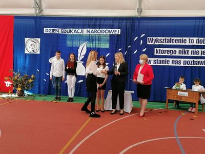 Dyrektor Szkoły Podstawowej wręcza nagrodę nauczycielce, obok pracownik administracyjny, w tle uczniowie placówki, którzy przygotowali akademię.