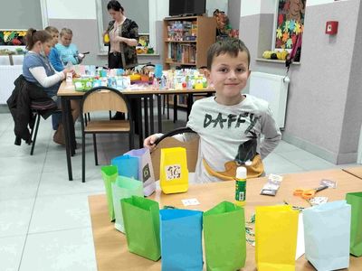 Chłopiec z PWD Kusice z przygotowanymi zadaniami do kalendarza, w tle inne dzieci i wychowawczyni.