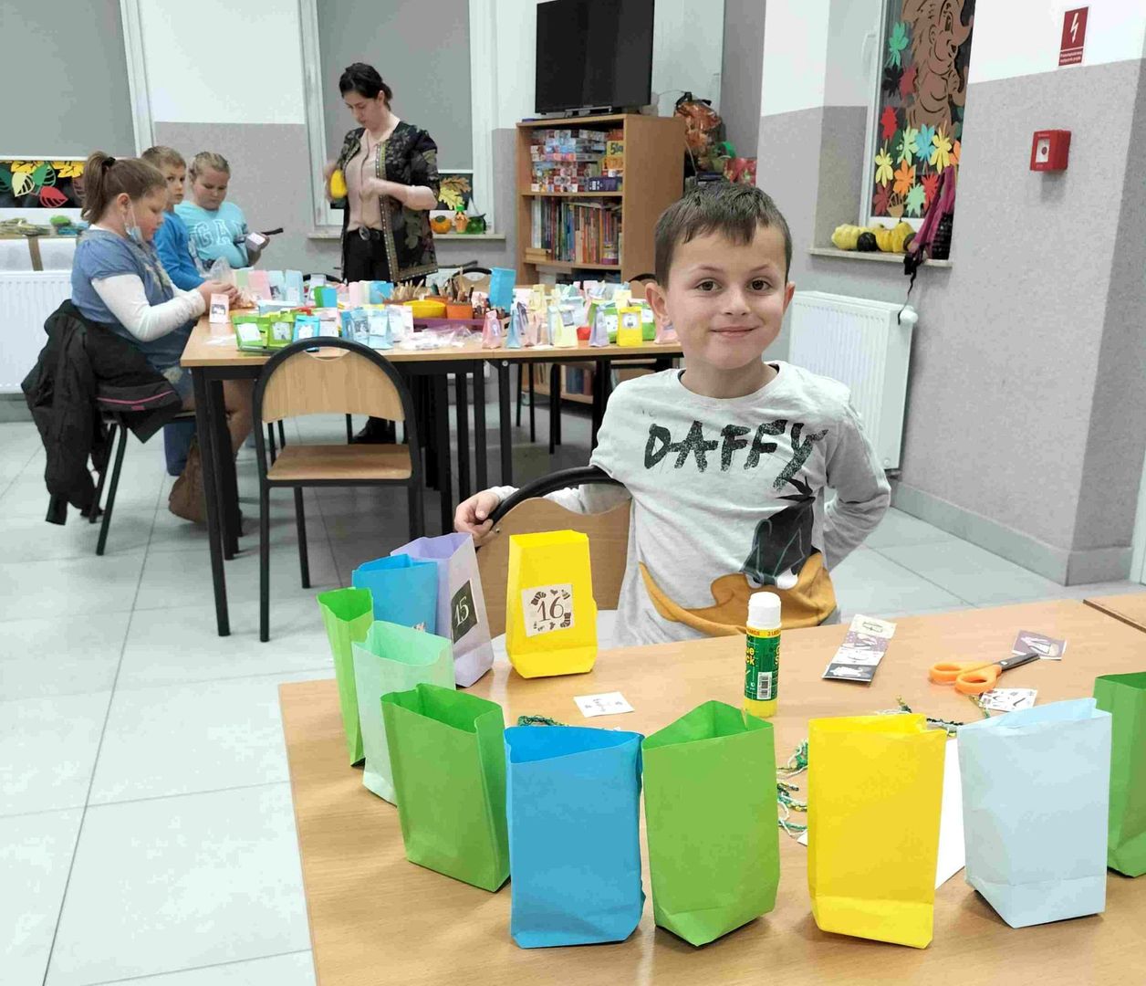 Chłopiec z PWD Kusice z przygotowanymi zadaniami do kalendarza, w tle inne dzieci i wychowawczyni.