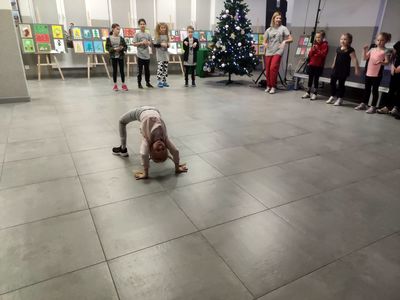 Pokaz breakdance, dziewczyna wykonuje mostek, w tle pozostałe dzieci kibicujące jej oraz trenerka.