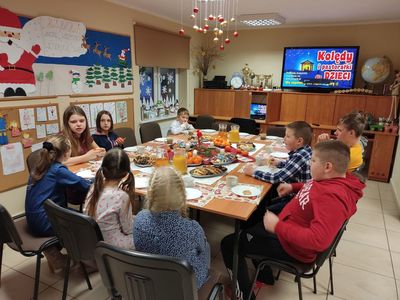 Wychowankowie z placówki wsparcia dziennego w Kosierzewie przy wspólnym wigilijnym stole, w tle relacja z Jasełek przygotowanych przez uczniów ZS w Ostrowcu.