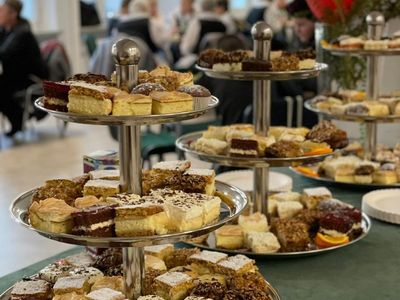 Patery z ciastami przygotowane w kawiarence, w tle osoby delektujące się wypiekami. 