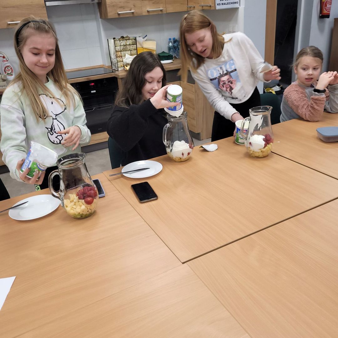 Dzieci ze Świetlicy Kultury w Malechowie podczas przygotowywania owocowych musów, w dzbankach owoce, dziewczynki wlewają do nich jogurt naturalny, pozostali obserwują.
