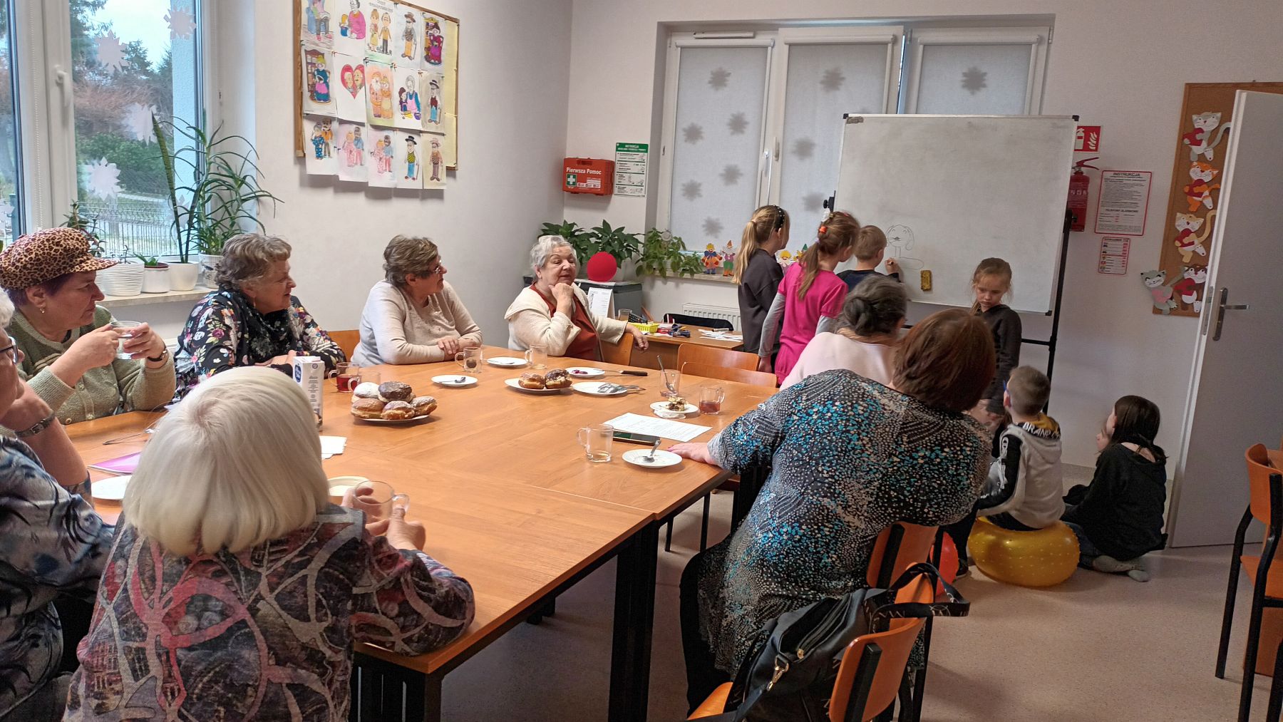 Dzieci z Placówki Wsparcia Dziennego w Ostrowcu wraz z seniorami podczas spożywania pączków.