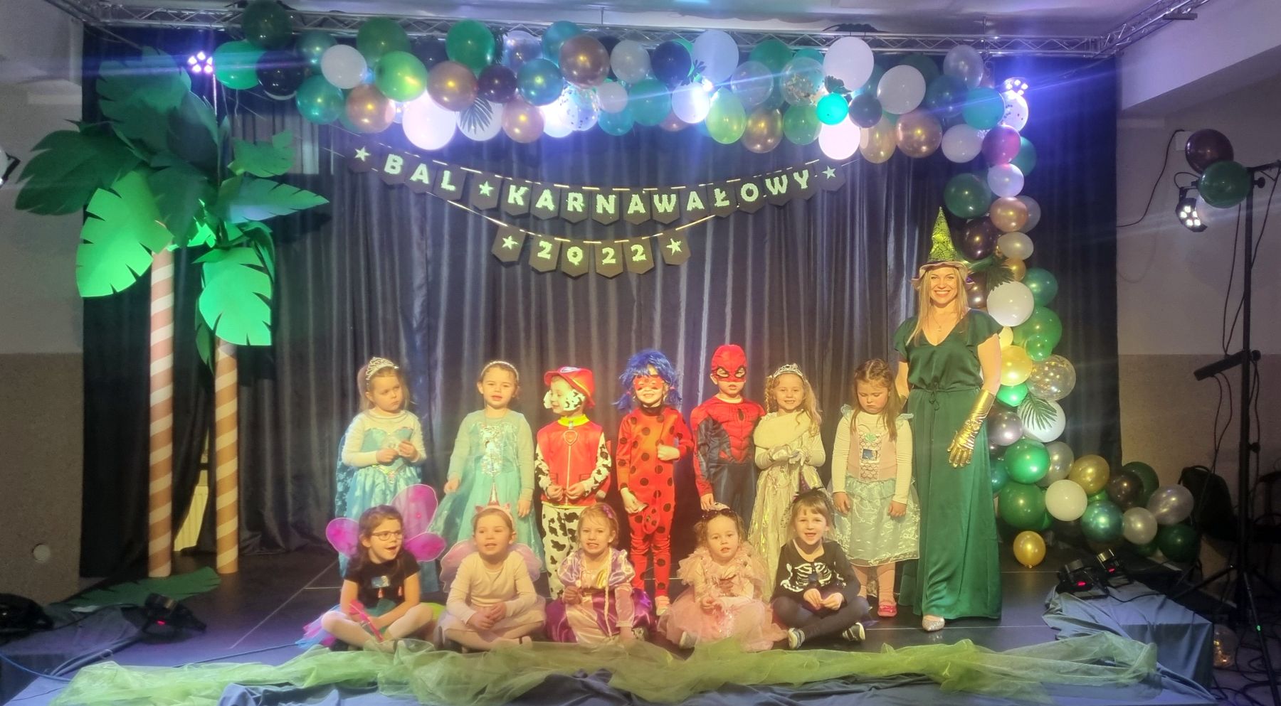 Grupa przedszkolaków na scenie w klimacie tropików wraz z wychowawczynią. Pozują do zdjęcia w przebraniach karnawałowych. 