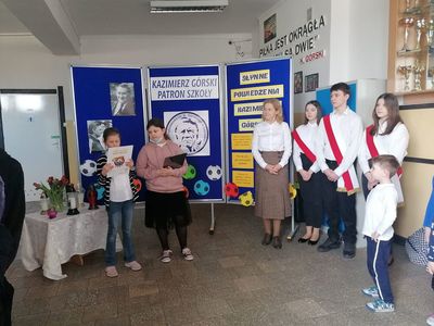 Dwie uczennice z SP Lejkowo prezentują informacje na temat patrona szkoły, obok dyrektor i poczet sztandarowy oraz uczniowie. 