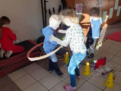Dziewczynka siedząca na matach gimnastycznych. Chłopiec kręcący hula-hop i dziewczynka pomagająca mu założyć je. Dziewczynka układająca pachołki.