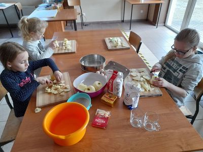 Trzy dziewczynki z Placówki Wsparcia Dziennego w Sulechowie kroją jabłka. Na stole widać produkty potrzebne do upieczenia ciasta - mąka, jajka, proszek do pieczenia, margaryna, cukier.