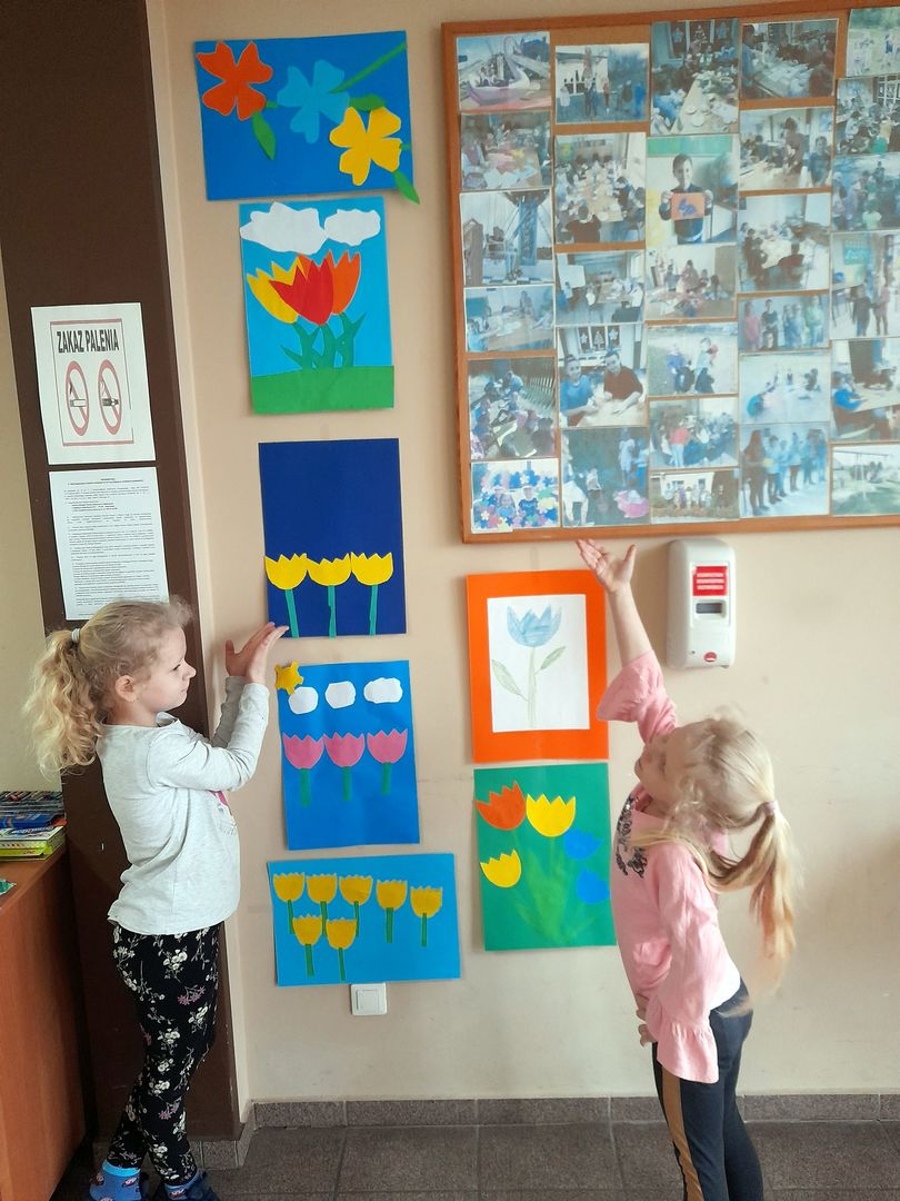 Dwie dziewczynki z Placówki Wsparcia Dziennego w Sulechowie prezentujące galerię prac przedstawiającą wiosenne kwiaty.