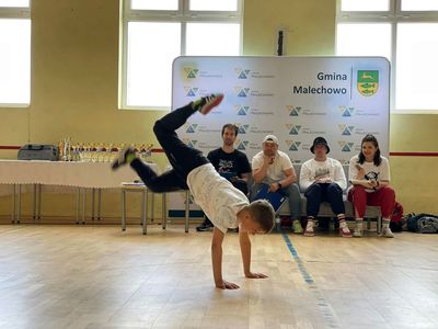 Zawodnik podczas pokazu na sali gimnastycznej w Malechowie, w tle sędziowie i prowadząca zawodów.