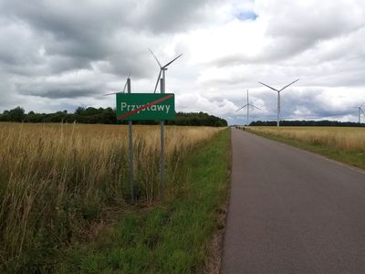 Znak drogowy z napisem Przystawy mówiący o kończącej się strefie wsi, w tle wiatraki i łany zbóż.  