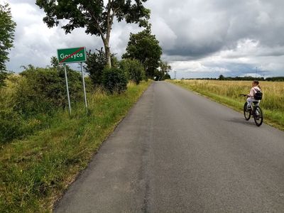 Znak drogowy z napisem Gorzyca mówiący o kończącej się strefie wsi, w tle dziewczynka na rowerze i i łany zbóż.  