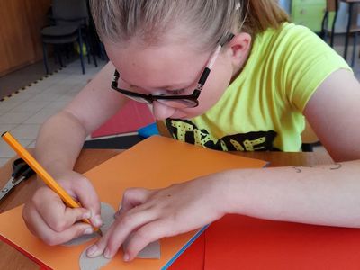 Dziewczynka z Placówki Wsparcia Dziennego w Sulechowie rysująca kwiat na kolorowym papierze przy użyciu szablonu.