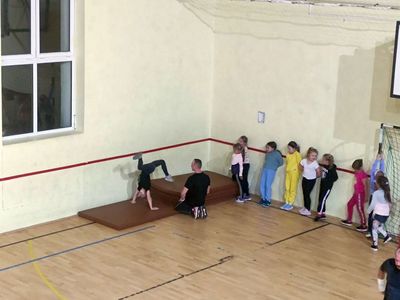 Dzieci z grupy zaawansowanej ćwiczą z trenerem w sali gimnastycznej. 