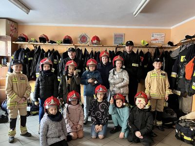 Dzieci w hełmach strażackich oraz w specjalistycznym ubraniu pozują do zdjęcia wraz ze strażakami.