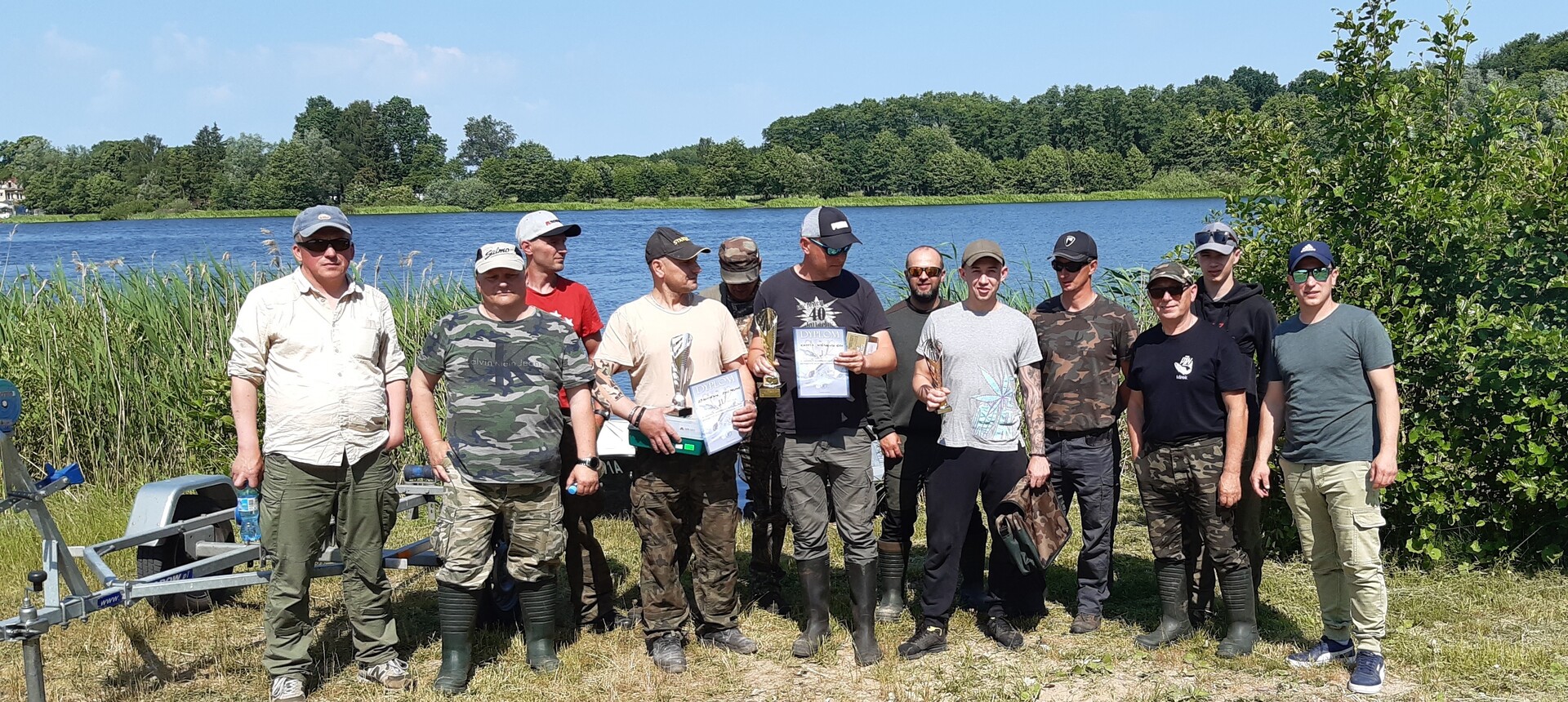 Uczestnicy zawodów wędkarskich z dyplomami i pucharami.