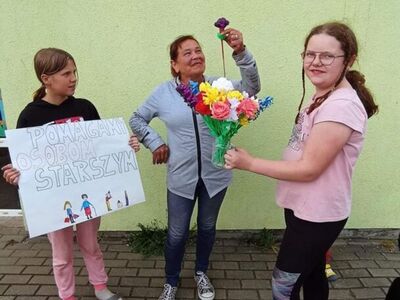 Wychowankowie z PWD z Lasek wręczają mieszkańcom Lasek kwiaty z krepiny i bibuły.