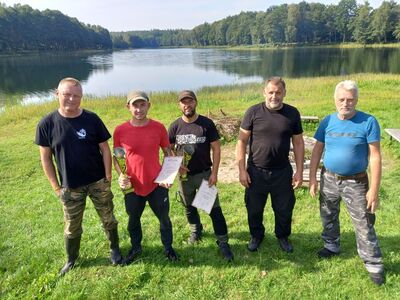 Nagrodzeni wędkarze z komisją sędziowską na tle jeziora.