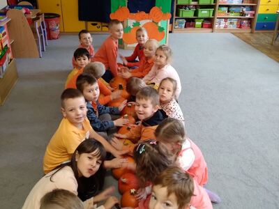 Dzieci w dwóch rzędach siedzą na dywanie - w środku dynia.