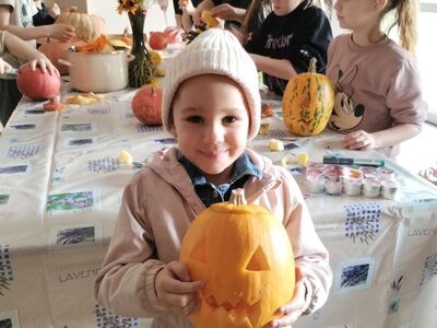 Dziewczynka prezentuje wykonany przez siebie lampion z dyni. W tle dzieci i rodzic podczas prac przy przygotowaniu lampionów.