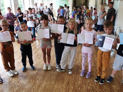 Na zdjęciach widać grupę dzieci w szkole. Dzieci trzymają w rękach otwarte zeszyty widoczną pieczątką, na której widnieje napis "Kordian". Dzieci ubrane w różnorodne ubrania, od sportowych do bardziej codziennych. W tle widać większą grupę dzieci oraz kilka osób dorosłych. Atmosfera wydaje się być radosna i swobodna, a dzieci prezentują swoje zeszyty z dumą.