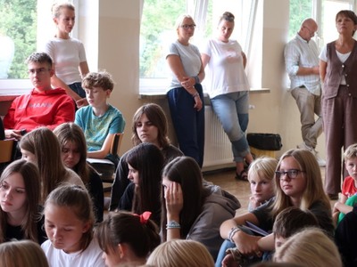 Na zdjęciach widać grupę dzieci w szkole. Dzieci trzymają w rękach otwarte zeszyty widoczną pieczątką, na której widnieje napis "Kordian". Dzieci ubrane w różnorodne ubrania, od sportowych do bardziej codziennych. W tle widać większą grupę dzieci oraz kilka osób dorosłych. Atmosfera wydaje się być radosna i swobodna, a dzieci prezentują swoje zeszyty z dumą.
