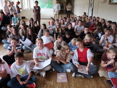 Na zdjęciach widać grupę dzieci w szkole. Dzieci trzymają w rękach otwarte zeszyty widoczną pieczątką, na której widnieje napis "Kordian". Dzieci ubrane w różnorodne ubrania, od sportowych do bardziej codziennych. W tle widać większą grupę dzieci oraz kilka osób dorosłych. Atmosfera wydaje się być radosna i swobodna, a dzieci prezentują swoje zeszyty z dumą.