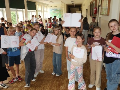 Na zdjęciach widać grupę dzieci w szkole. Dzieci trzymają w rękach otwarte zeszyty widoczną pieczątką, na której widnieje napis "Kordian". Dzieci ubrane w różnorodne ubrania, od sportowych do bardziej codziennych. W tle widać większą grupę dzieci oraz kilka osób dorosłych. Atmosfera wydaje się być radosna i swobodna, a dzieci prezentują swoje zeszyty z dumą.