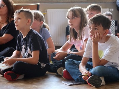 Na zdjęciach widać grupę dzieci w szkole. Dzieci trzymają w rękach otwarte zeszyty widoczną pieczątką, na której widnieje napis "Kordian". Dzieci ubrane w różnorodne ubrania, od sportowych do bardziej codziennych. W tle widać większą grupę dzieci oraz kilka osób dorosłych. Atmosfera wydaje się być radosna i swobodna, a dzieci prezentują swoje zeszyty z dumą.