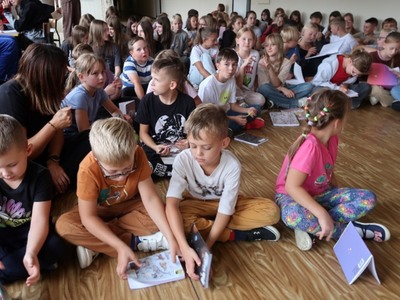 Na zdjęciach widać grupę dzieci w szkole. Dzieci trzymają w rękach otwarte zeszyty widoczną pieczątką, na której widnieje napis "Kordian". Dzieci ubrane w różnorodne ubrania, od sportowych do bardziej codziennych. W tle widać większą grupę dzieci oraz kilka osób dorosłych. Atmosfera wydaje się być radosna i swobodna, a dzieci prezentują swoje zeszyty z dumą.
