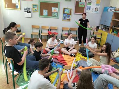 Na zdjęciu młodzież uczestniczy w warsztatach z modelowania balonów, siedzą po turecku na podłodze i modelują balony.