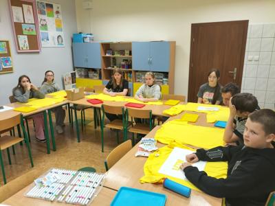 Nas zdjęciu młodzież siedzi przy stoliku i malują farbami żółte, projektowe koszulki.