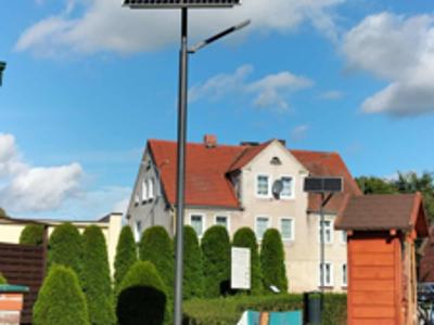 Na zdjęciu lampa solarna zamontowana na terenie wokół ośrodka kultury, na terenie zielonym znajduje się lampa, ławeczka, plenerowa biblioteczka, krzewy.